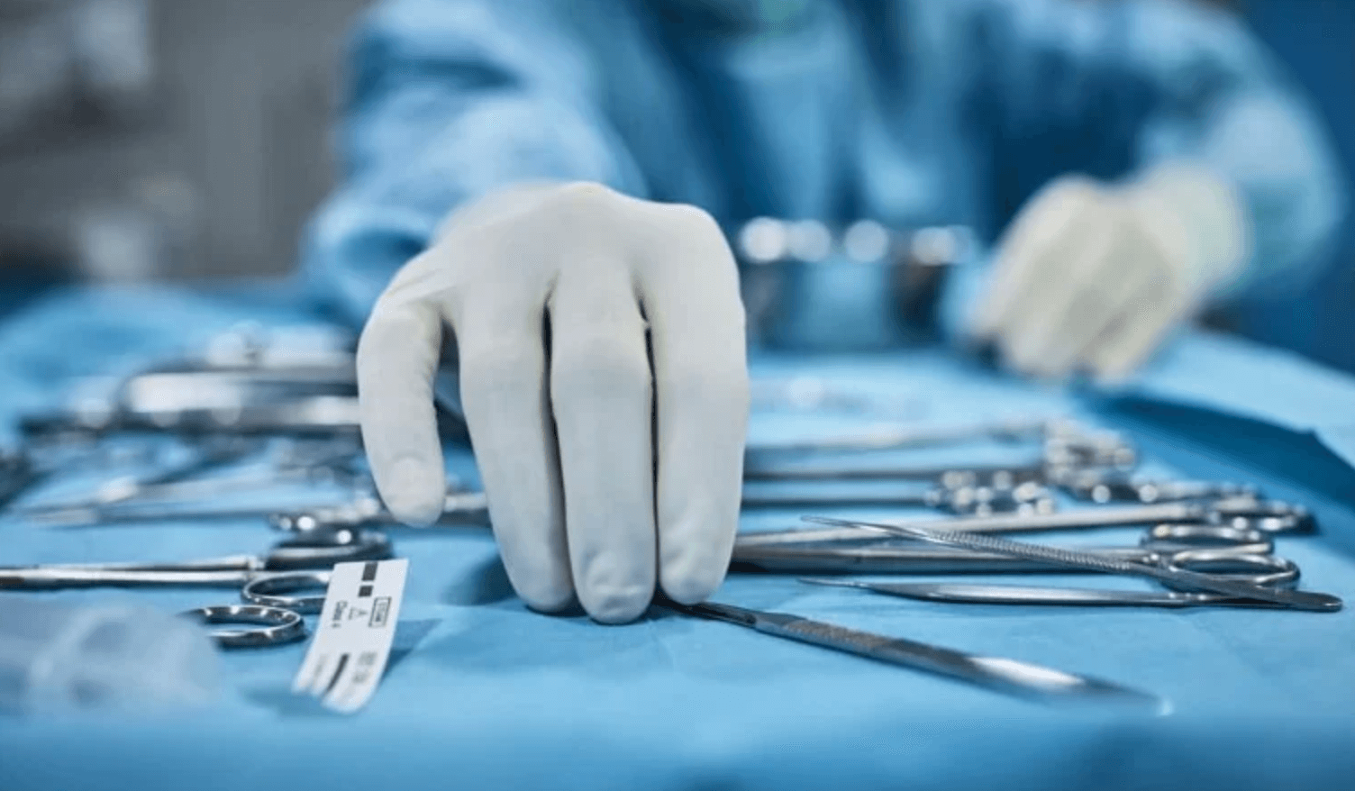 A gloved hand reaches for a surgical instrument on a table covered with various metal tools. The person is wearing blue surgical attire, following ANSI/AAMI ST108:2023 Standards, and the background is slightly out of focus, suggesting a medical or surgical setting.